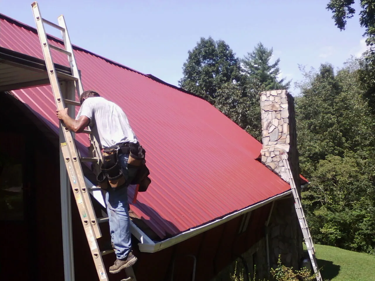 Roofer on a ladder beside a red metal roof and stone chimney — Soffit & Fascia Repair by Roofing Brothers in House Springs
